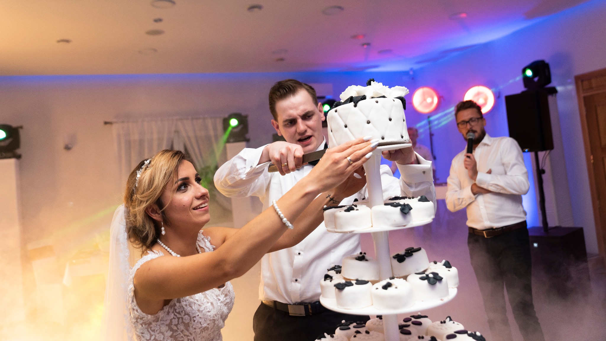 Para młoda kroi wielopiętrowy tort weselny, czarno-biały z dekoracjami kwiatowymi, w tle konferansjer z mikrofonem i oświetlenie sceniczne.