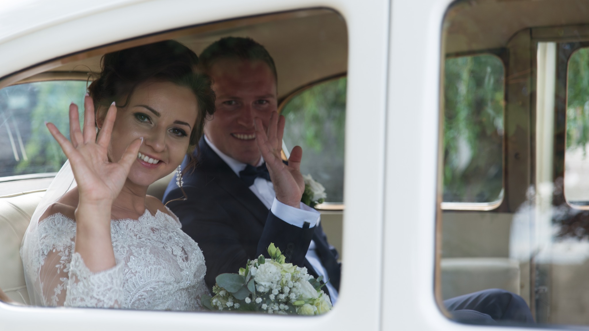 Uśmiechnięta para młoda w klasycznym białym samochodzie, machająca przez okno, bukiet ślubny na kolanach panny młodej, ciemny garnitur pana młodego z muchą.