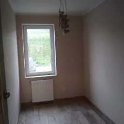 Pusty pokój po remoncie z drewnopodobną podłogą, świeżo pomalowanymi ścianami w odcieniach beżu, białym oknem z widokiem na zieleń i grzejnikiem pod nim, oraz oryginalną lampą wiszącą z tektury.