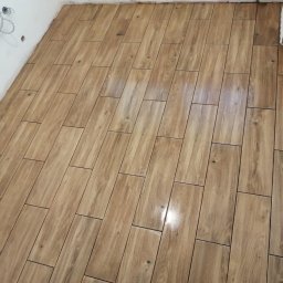 DAMEX-BUD - Ułożona podłoga z płytek imitujących drewno w łazience, widoczne niedokończone instalacje na ścianie i stary grzejnik.