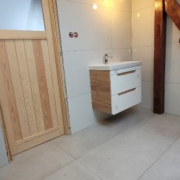 DAMEX-BUD - Wnętrze łazienki w trakcie wykańczania, widoczna drewniana rama drzwi, białe płytki na ścianach, podwieszana szafka z umywalką oraz drewniane belki stropowe z żarówkami na kablach.