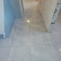 DAMEX-BUD - Błyszcząca, jasna podłoga z płytek w trakcie remontu, widoczne ślady po pracach na ścianach i futrynach drzwi, para butów w dolnej części kadru.