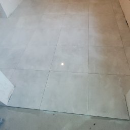 DAMEX-BUD - Świeżo ułożone, jasnoszare płytki podłogowe w surowym stanie łazienki, widoczne fragmenty nieotynkowanych ścian i przyłącza instalacyjne.