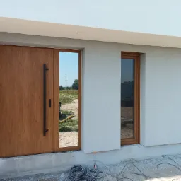 Wejście do domu w trakcie wykańczania: drewniane drzwi z długim, czarnym uchwytem, okno z brązową ramą, biała elewacja o fakturze baranka, widoczne kable na ziemi.