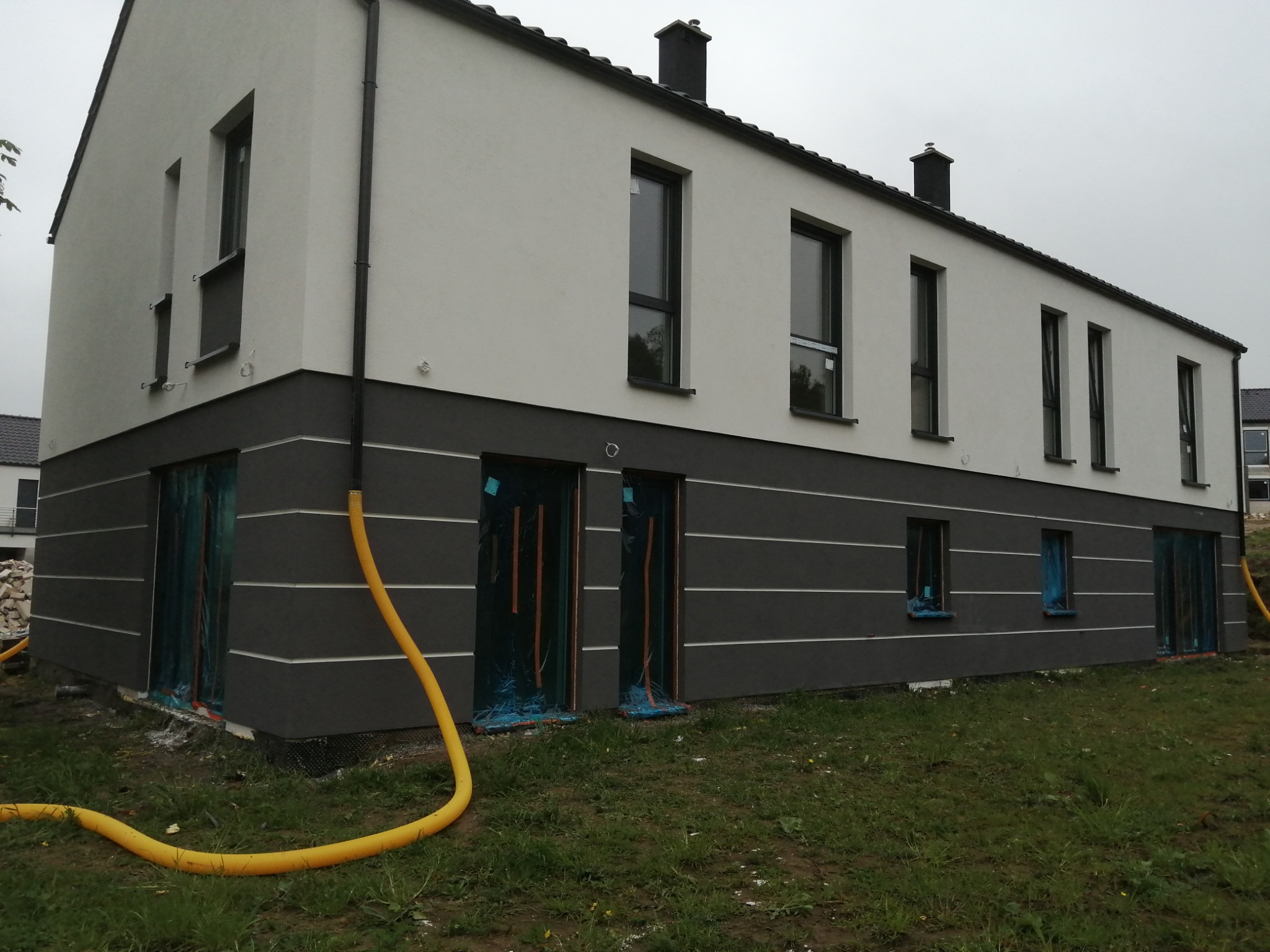 Nowoczesny, dwukondygnacyjny dom z elewacją w dwóch odcieniach szarości i białym wykończeniem, z oknami zabezpieczonymi folią, z widocznym żółtym wężem na trawniku, sugerującym trwające prace.