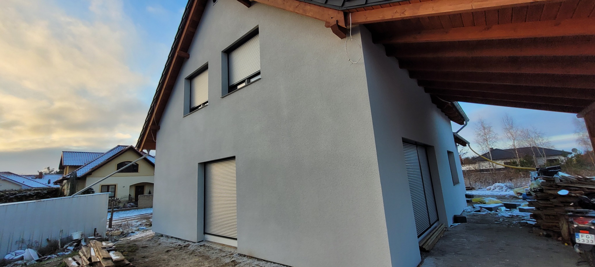 Nowo otynkowany dom jednorodzinny z szarym tynkiem i roletami zewnętrznymi, widok z boku z elementami placu budowy i stertą drewna pod zadaszeniem.