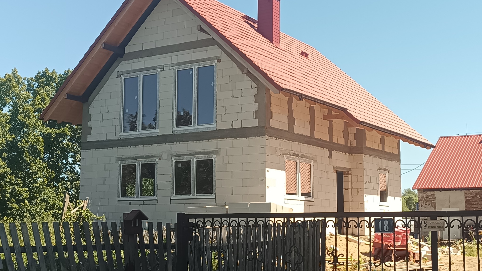 Dwukondygnacyjny dom w trakcie budowy z czerwonym dachem, widoczny zza ogrodzenia. Białe bloczki, okna wstawione, brak elewacji. Częściowo widoczne drzewa w tle.