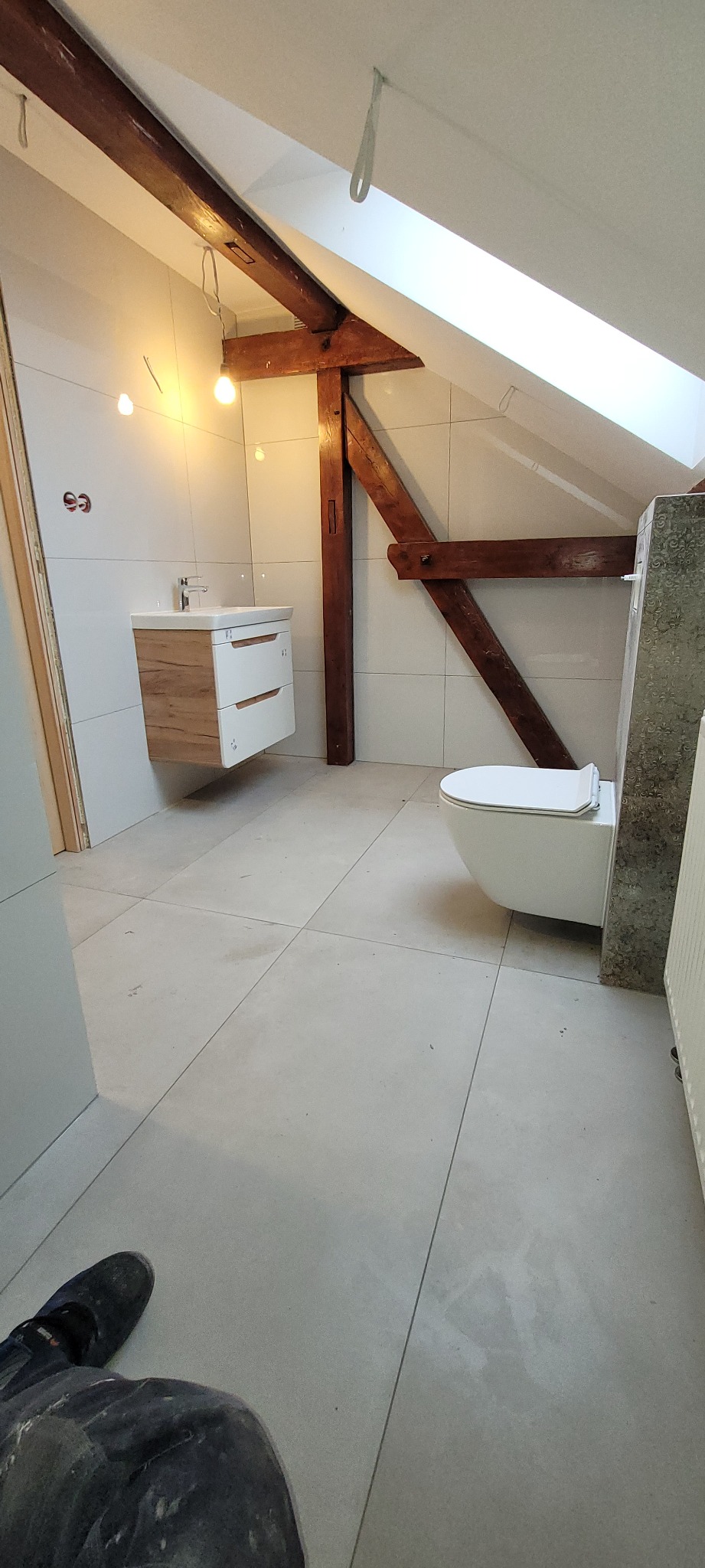 Łazienka na poddaszu ze skośnym sufitem, drewnianymi belkami konstrukcyjnymi, umywalką z szafką, podwieszaną toaletą i jasnymi płytkami podłogowymi oraz ściennymi.