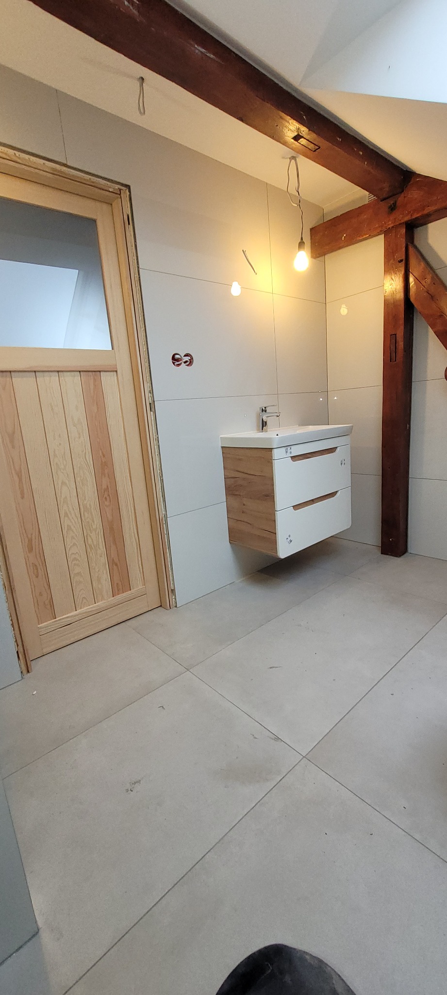 Wnętrze łazienki w trakcie wykańczania, widoczna drewniana rama drzwi, białe płytki na ścianach, podwieszana szafka z umywalką oraz drewniane belki stropowe z żarówkami na kablach.