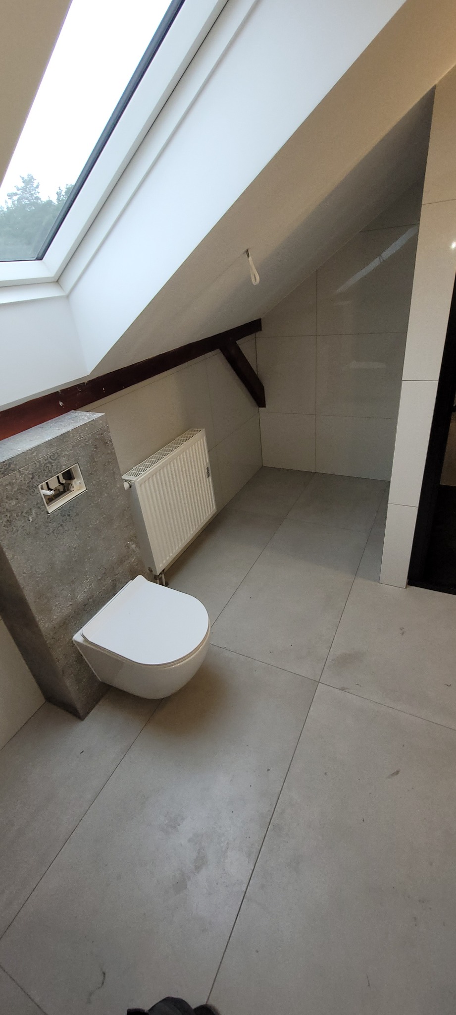 Nowoczesna, jasna łazienka na poddaszu w Gorzowie Wielkopolskim z wiszącą toaletą i grzejnikiem, wykończona szarymi płytkami wielkoformatowymi i oknem dachowym. Widoczna drewniana belka konstrukcyjna.