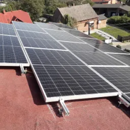 Panele słoneczne zamontowane na czerwonym dachu, widoczne aluminiowe konstrukcje mocujące i uziemienie, w tle zabudowa jednorodzinna.