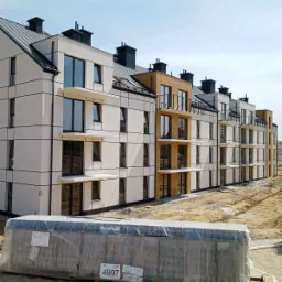 Nowoczesny budynek wielorodzinny z wykończoną elewacją w kolorze białym i fragmentami w odcieniu drewna, z balkonami i czarnymi ramami okiennymi, na pierwszym planie paleta z materiałami...