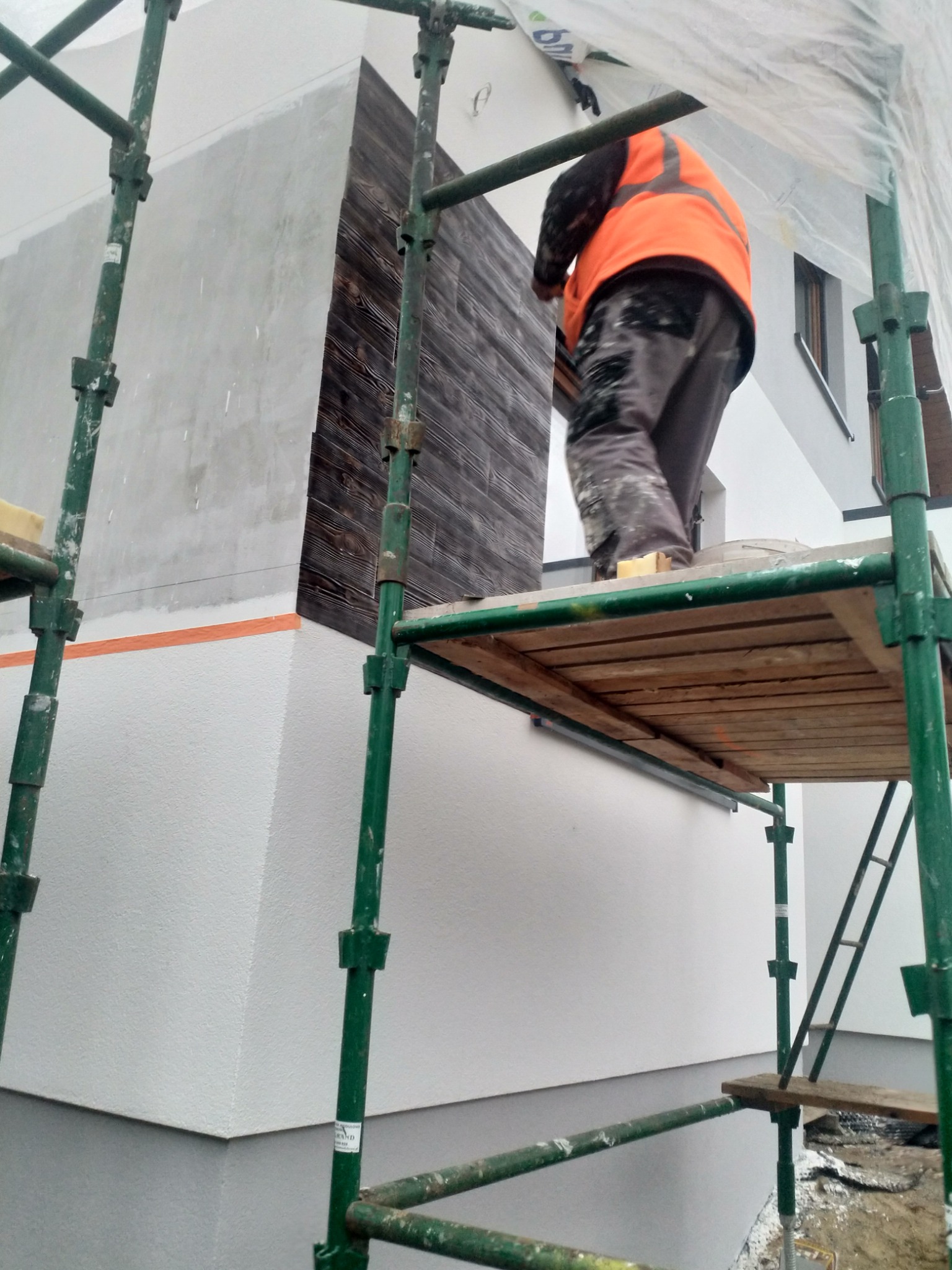 Pracownik w pomarańczowej kamizelce na rusztowaniu podczas montażu paneli imitujących drewno na elewacji budynku, widoczne fragmenty ocieplonej i otynkowanej ściany.