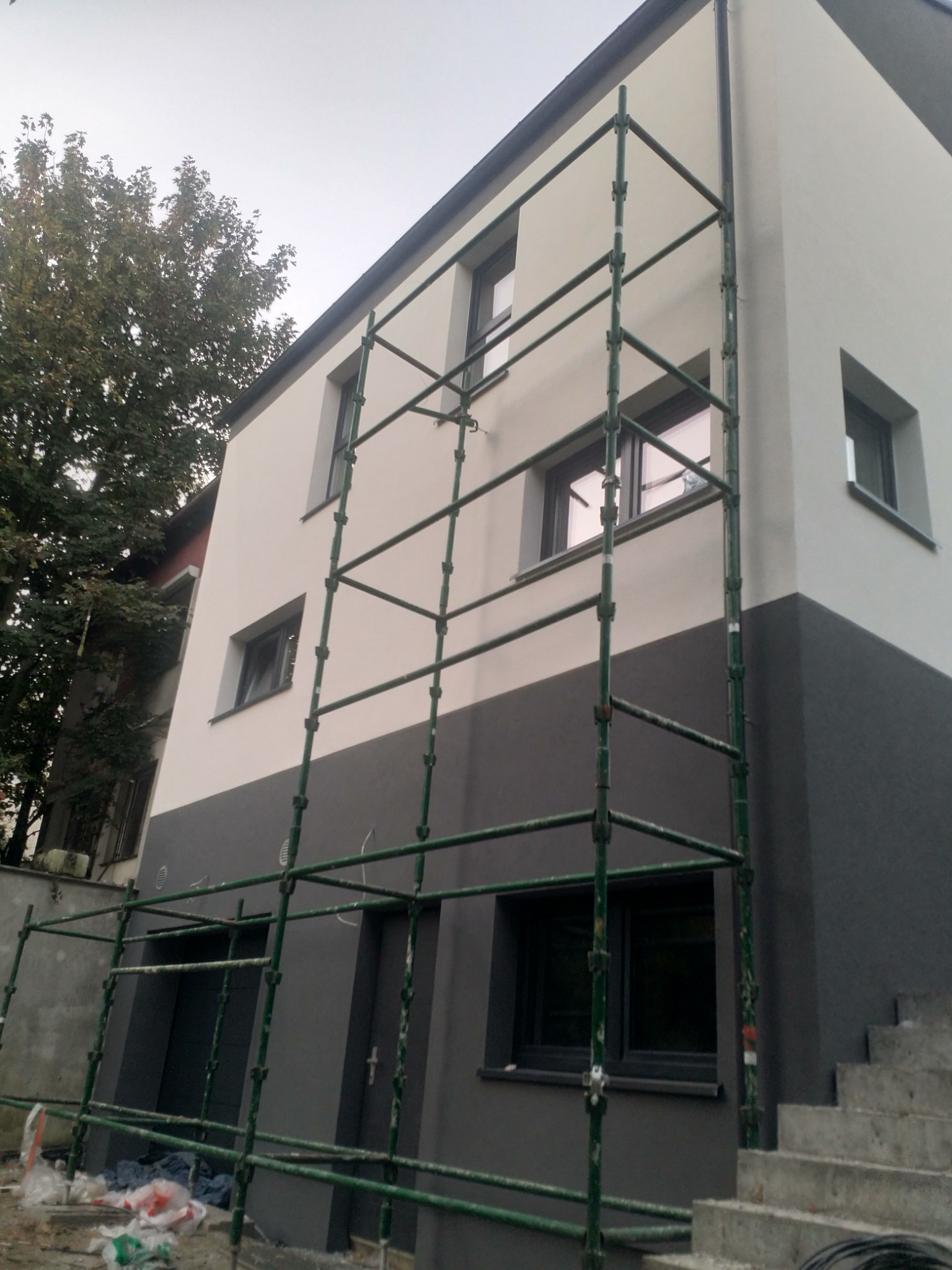 Dwukolorowa elewacja budynku w trakcie prac, z widocznym rusztowaniem i schodami wejściowymi.