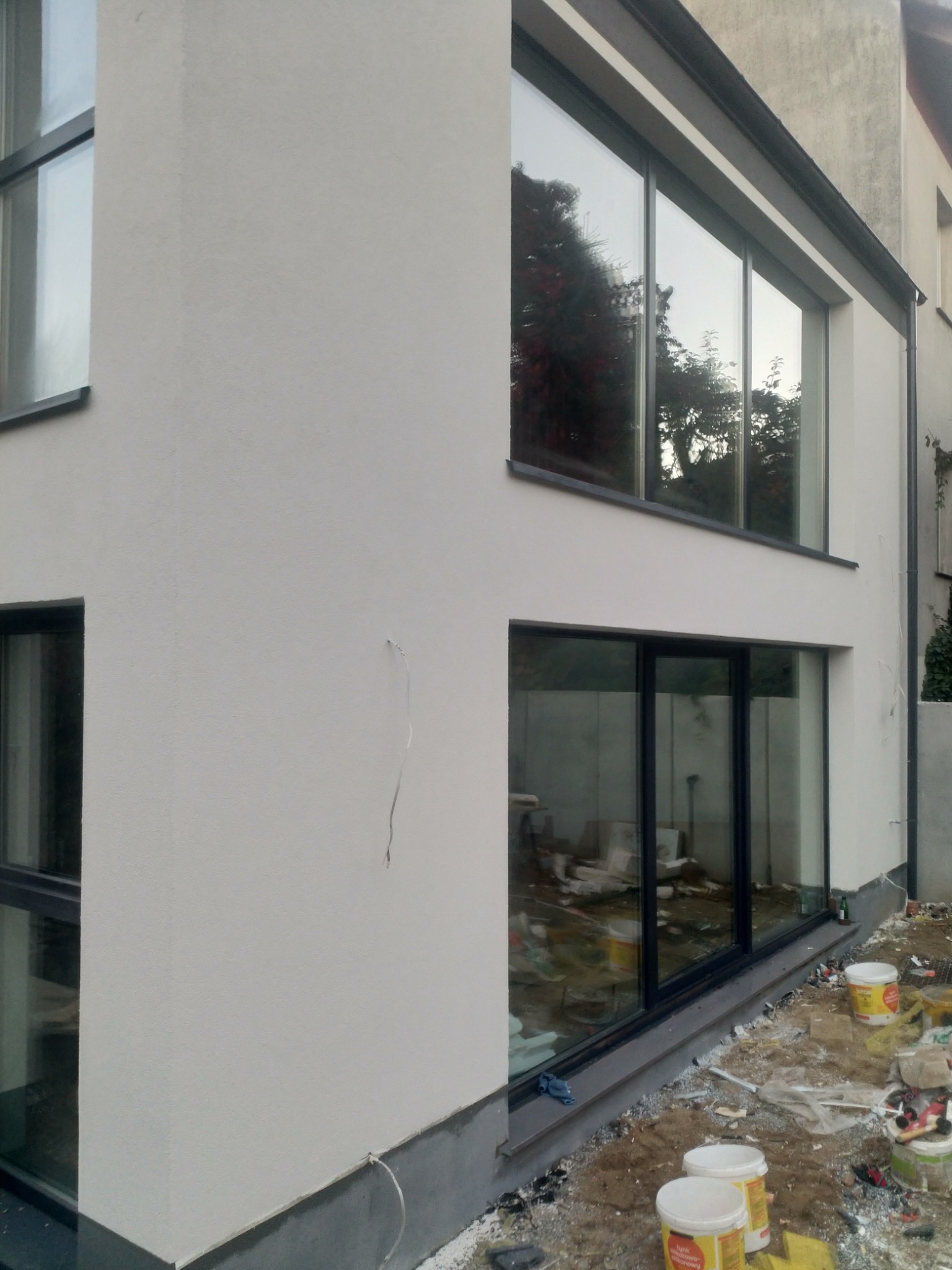 Fragment nowo otynkowanej elewacji domu z dużymi oknami, widoczne wiadra po farbie i resztki materiałów budowlanych na ziemi.
