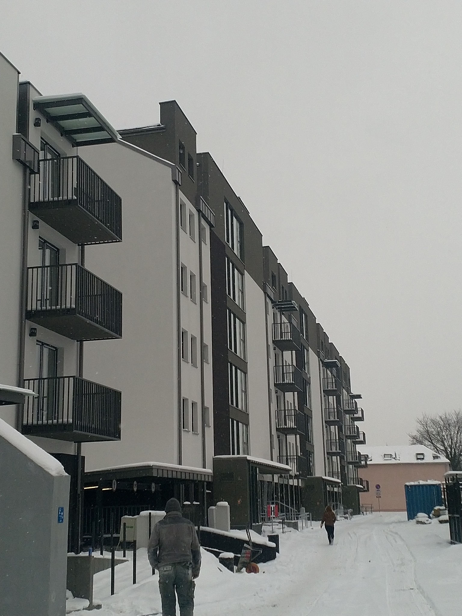 Nowoczesny budynek wielorodzinny z balkonami w zimowej scenerii, widoczni przechodnie i świeży śnieg.