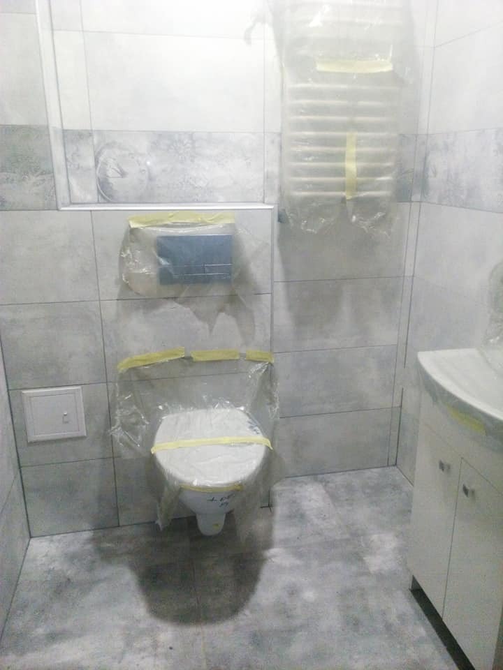 Przygotowane do remontu pomieszczenie łazienki: toaleta, grzejnik i spłuczka zabezpieczone folią i taśmą malarską, płytki ceramiczne na ścianach i podłodze, umywalka z szafką.