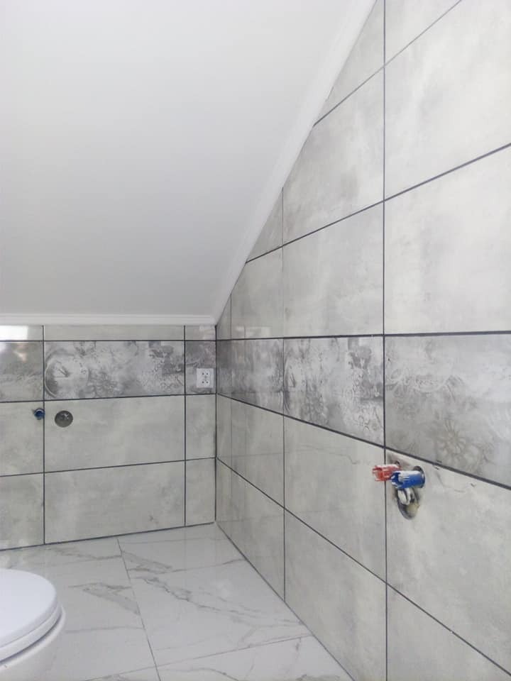 Wykończona łazienka z szarymi, kwadratowymi płytkami imitującymi beton, marmurową podłogą i wystającymi rurami instalacji wodnej.