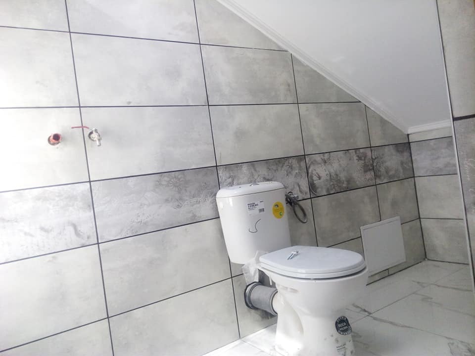 Nowa toaleta z białą ceramiką sanitarną na tle szarych, kwadratowych płytek z wzorem marmuru, widoczne podejścia instalacyjne do baterii.