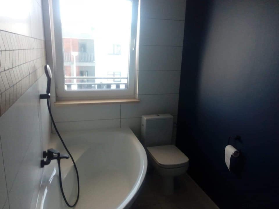 Nowoczesna łazienka z białą wanną, toaletą i ciemną ścianą, widok z okna na budynek mieszkalny.