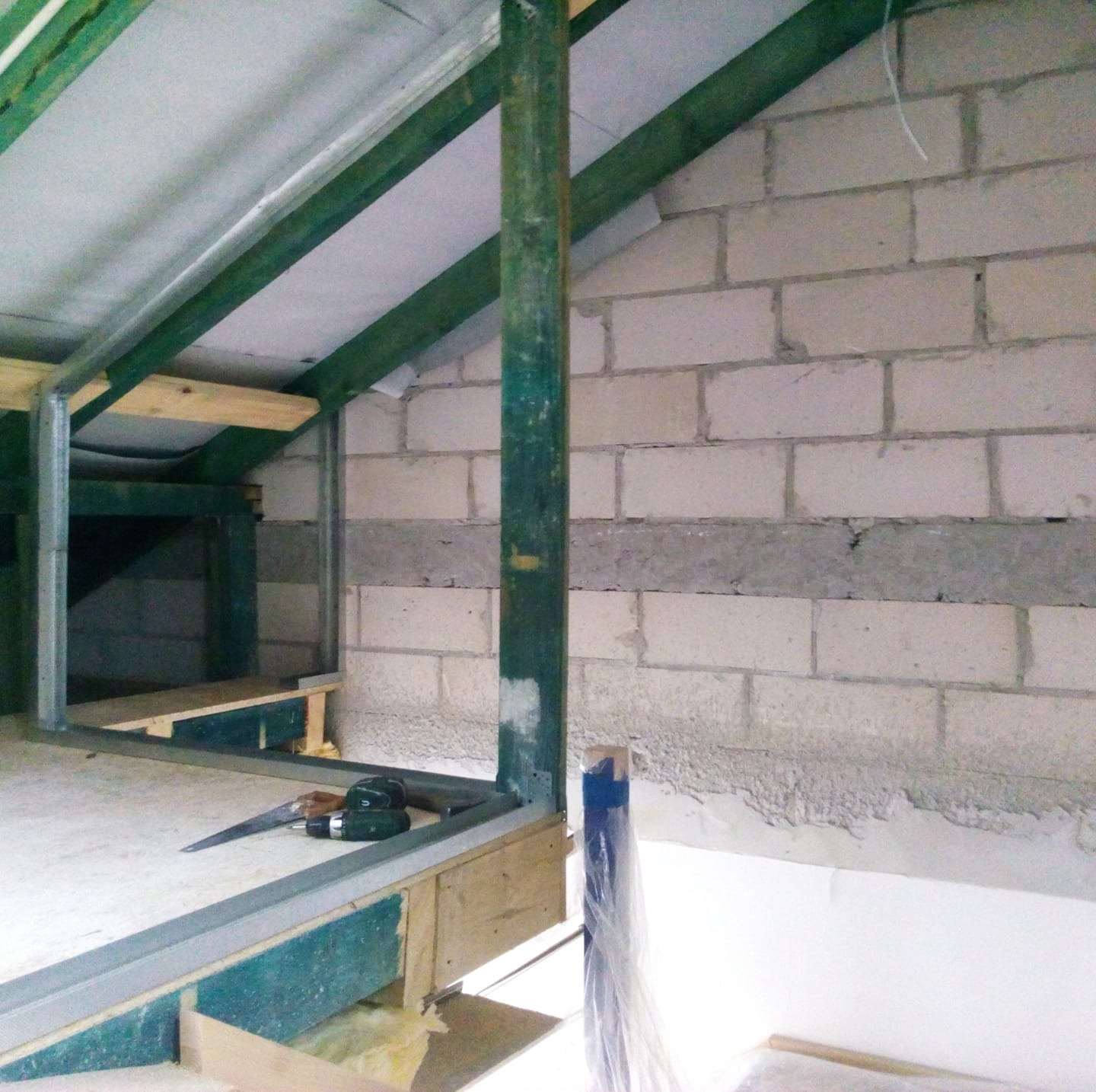 Wnętrze poddasza w trakcie adaptacji: surowa ściana z białego bloczka, metalowa konstrukcja szkieletowa obudowy, narzędzia (wkrętarka, nóż) na podłodze z płyty OSB, widoczna izolacja z wełny...