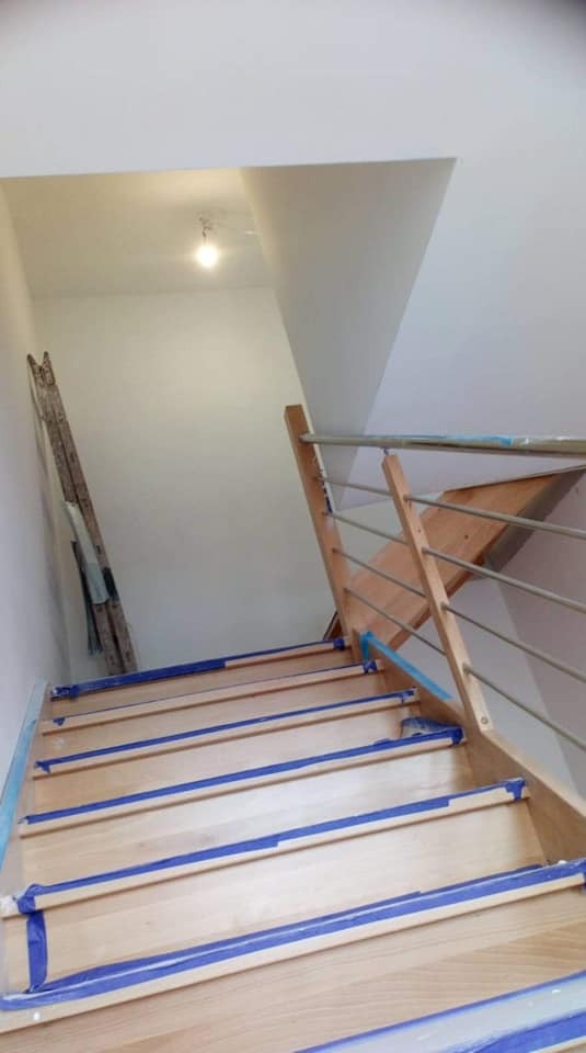 Schody drewniane zabezpieczone niebieską taśmą malarską, ściana świeżo pomalowana na biało, w tle drabina oparta o ścianę.