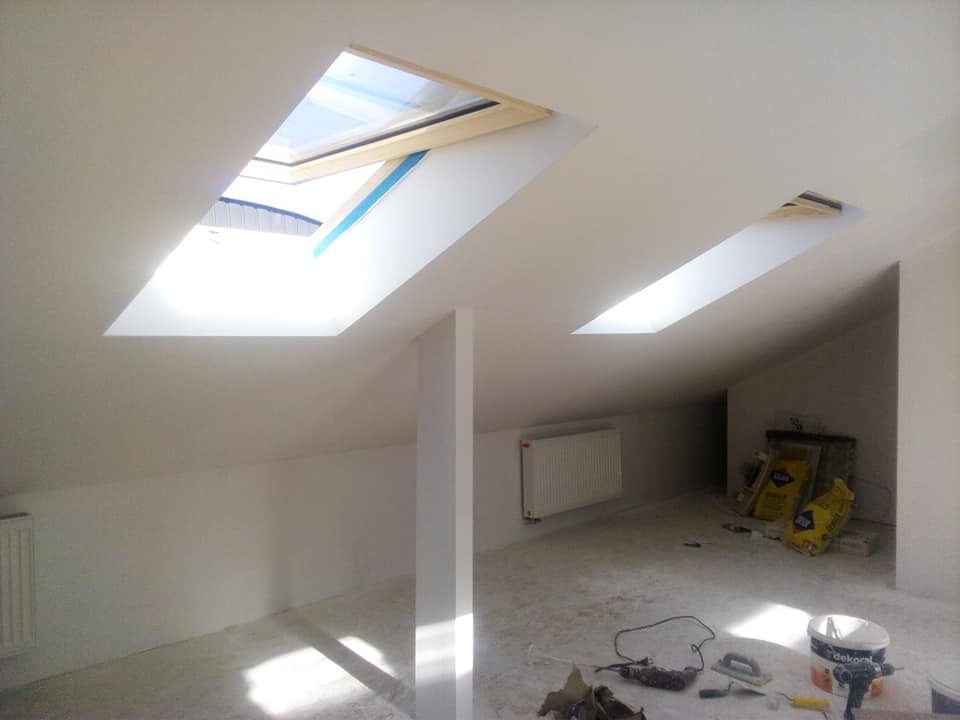 Poddasze po remoncie, widoczne dwa okna dachowe wpuszczające jasne światło, białe ściany i sufit, grzejnik oraz narzędzia i materiały budowlane na podłodze.