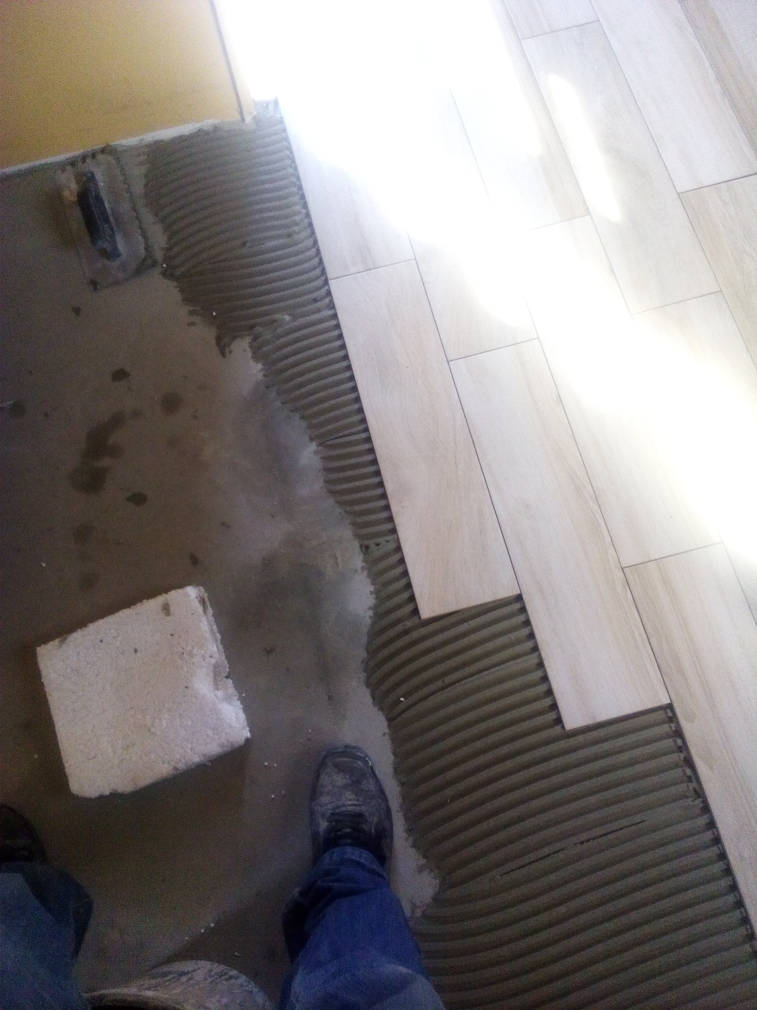 Układanie podłogi z płytek imitujących drewno, widoczna warstwa kleju z charakterystycznymi rowkami, fragment stopy w roboczym obuwiu i spodniach jeansowych, oraz zaprawa murarska.