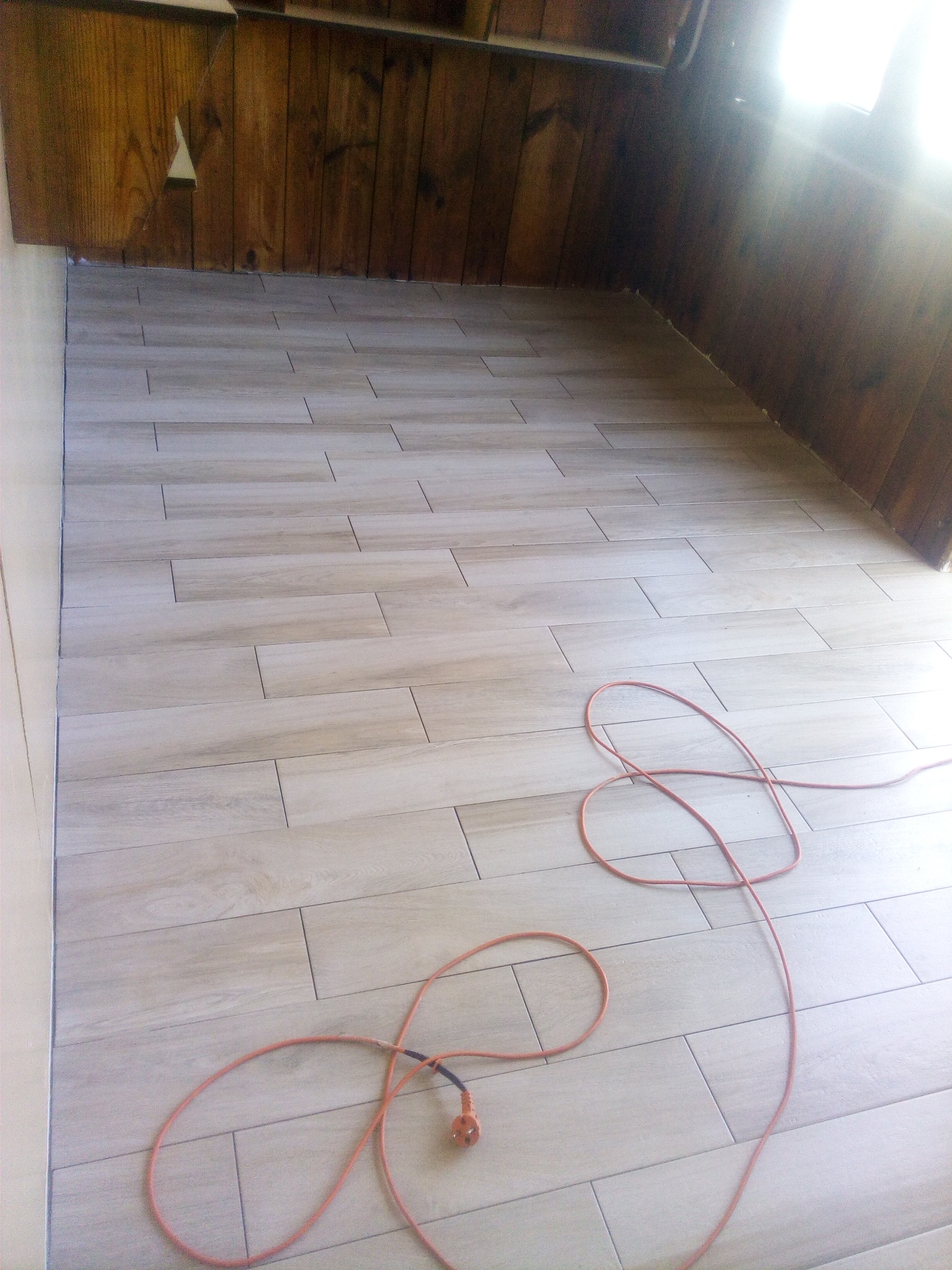 Ułożone płytki podłogowe imitujące drewno w pomieszczeniu z boazerią, widoczny pomarańczowy przedłużacz.