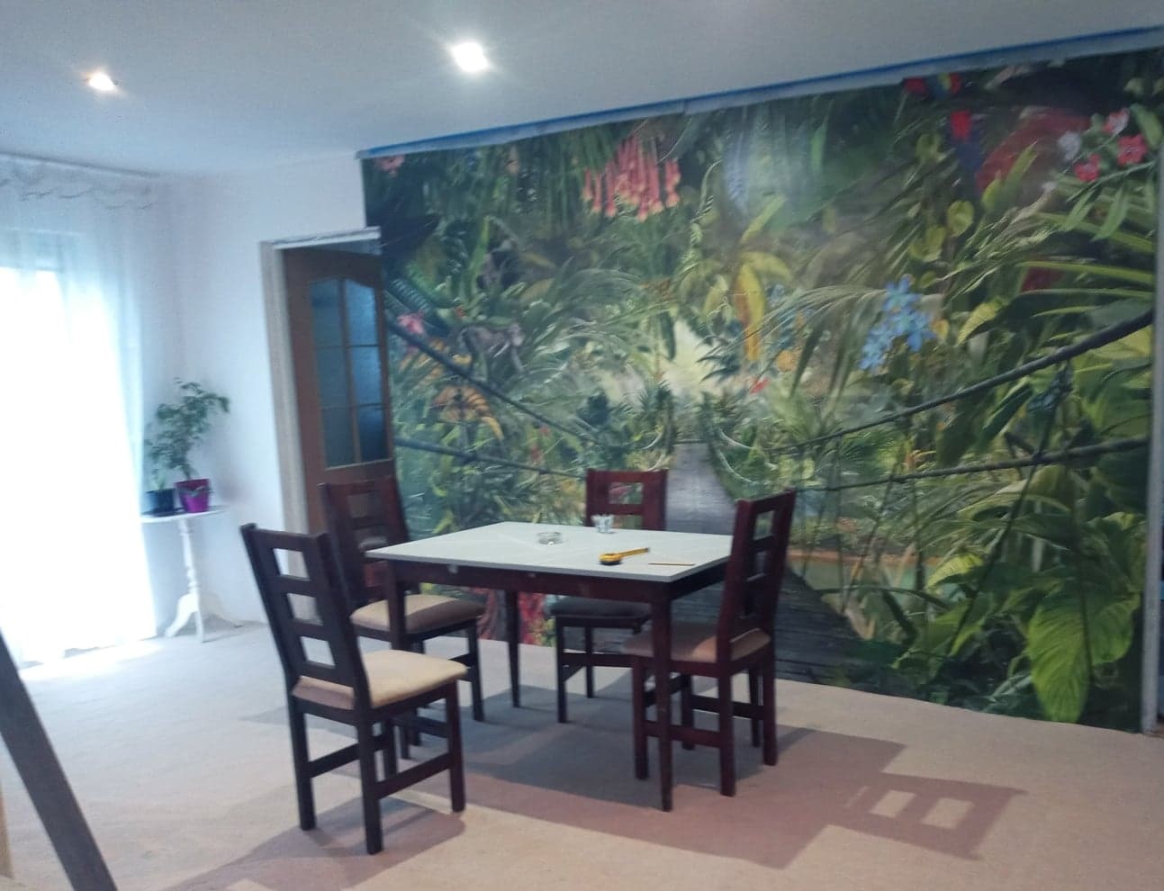 Pokój z fototapetą przedstawiającą tropikalny las, stół z czterema krzesłami, roślina doniczkowa przy oknie.