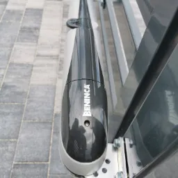 Szary siłownik bramy marki Beninca zamontowany na metalowej konstrukcji bramy przesuwnej, widoczne śruby mocujące i kostka brukowa w tle.