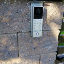 Domofon z kamerą zamontowany na kamiennym słupku ogrodzenia, widoczne przyciski numeryczne i logo producenta ORNO.