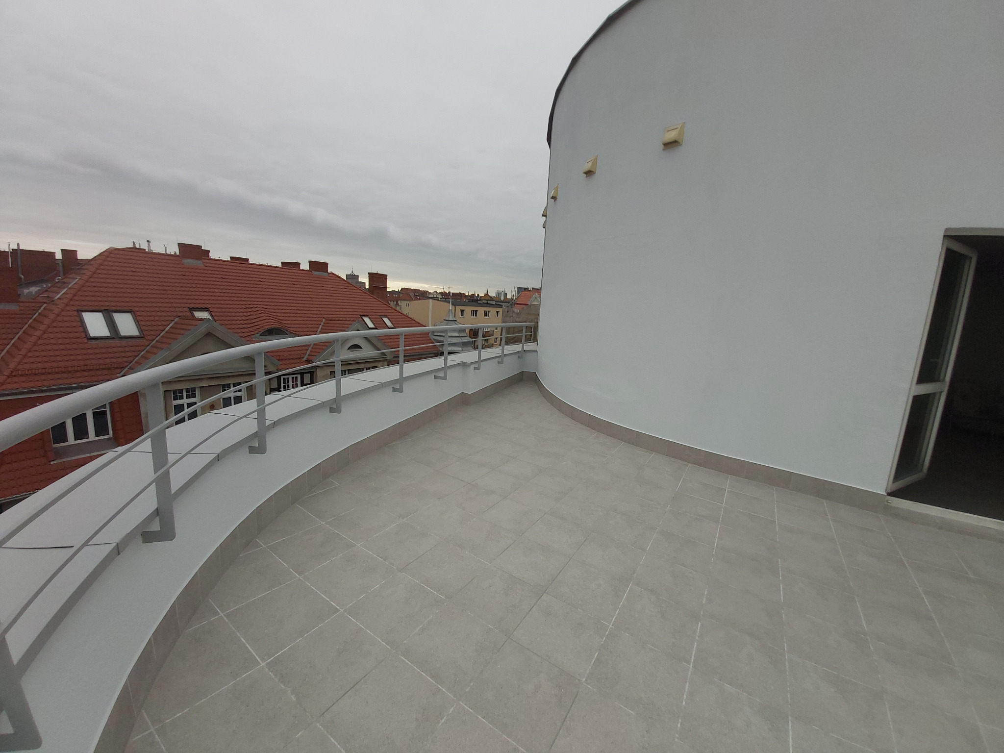 Wykończony balkon z szarymi płytkami i metalową balustradą na tle miejskiego krajobrazu z czerwonymi dachami.