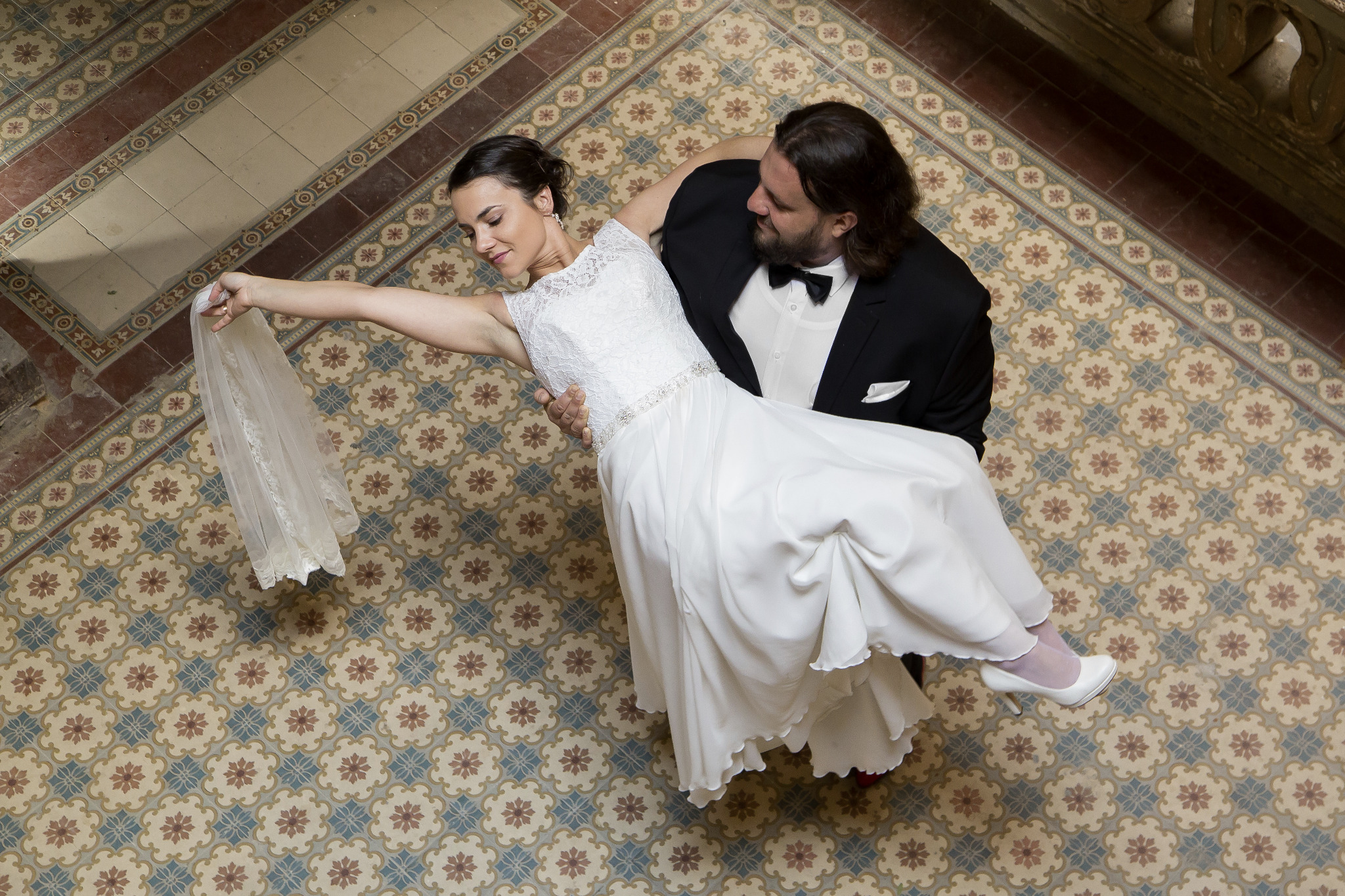 Para młoda: Panna Młoda w białej sukni ślubnej z koronkową górą i welonem trzymanym w ręce, unoszona przez Pana Młodego w czarnym smokingu z muchą, na tle wzorzystej podłogi.