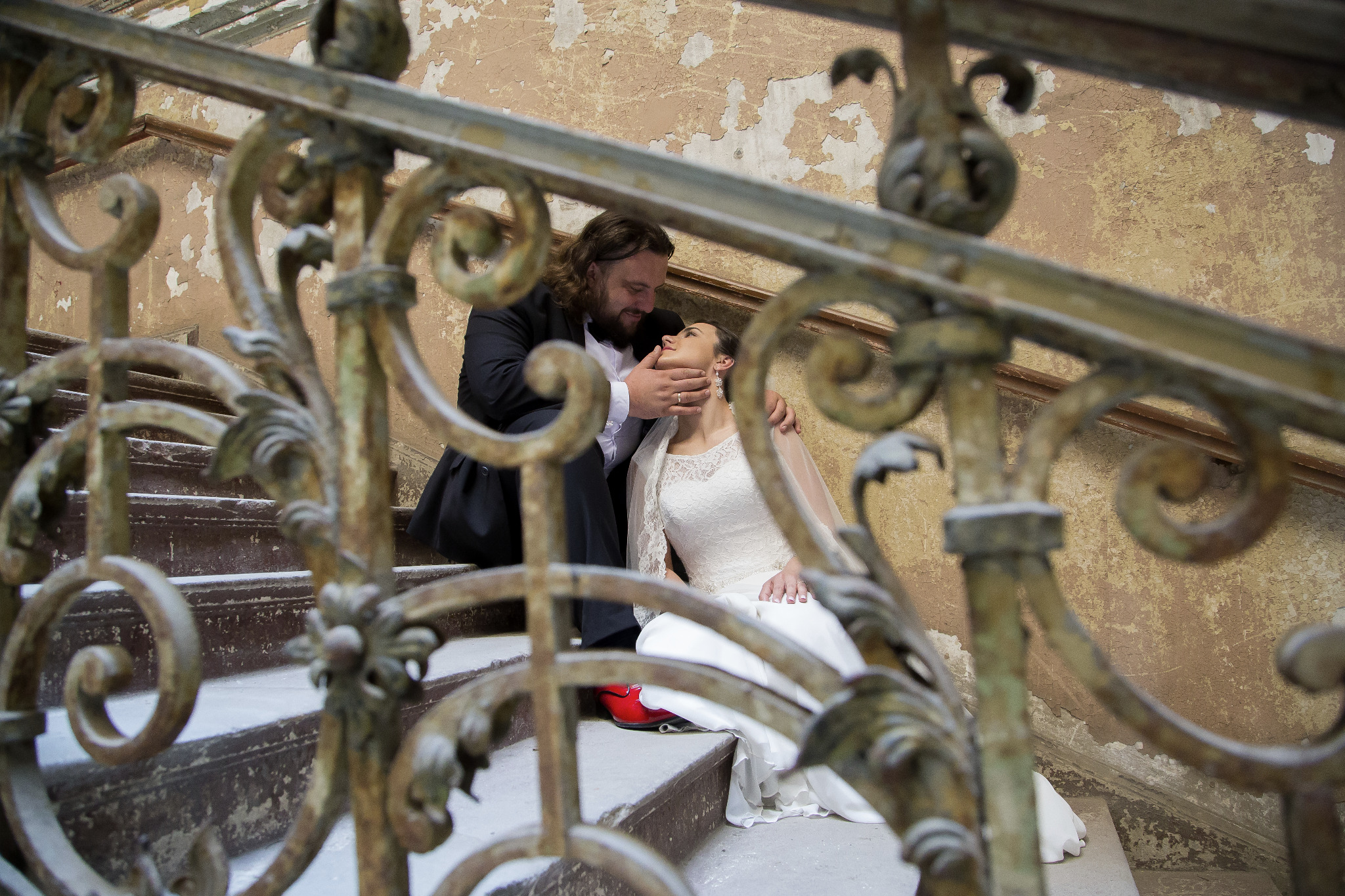 Para młoda siedzi na starych schodach z ozdobną, metalową balustradą; panna młoda w białej sukni i czerwonych butach, pan młody w czarnym garniturze.