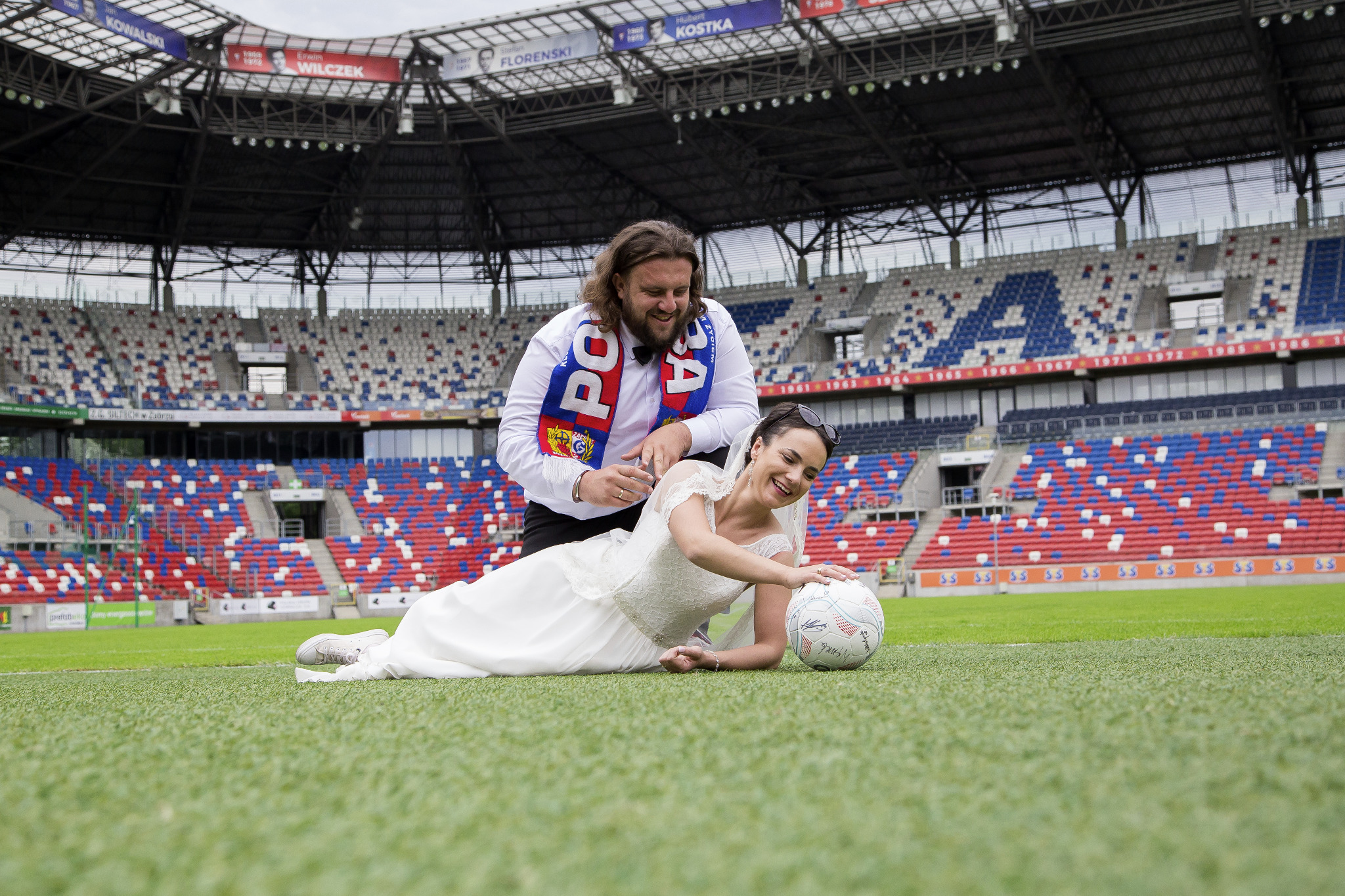 Uśmiechnięta panna młoda w sukni ślubnej i białych trampkach leży na murawie stadionu piłkarskiego, opierając ręce na piłce. Obok stoi pan młody z szalikiem kibica. W tle widoczne trybuny stadionu.