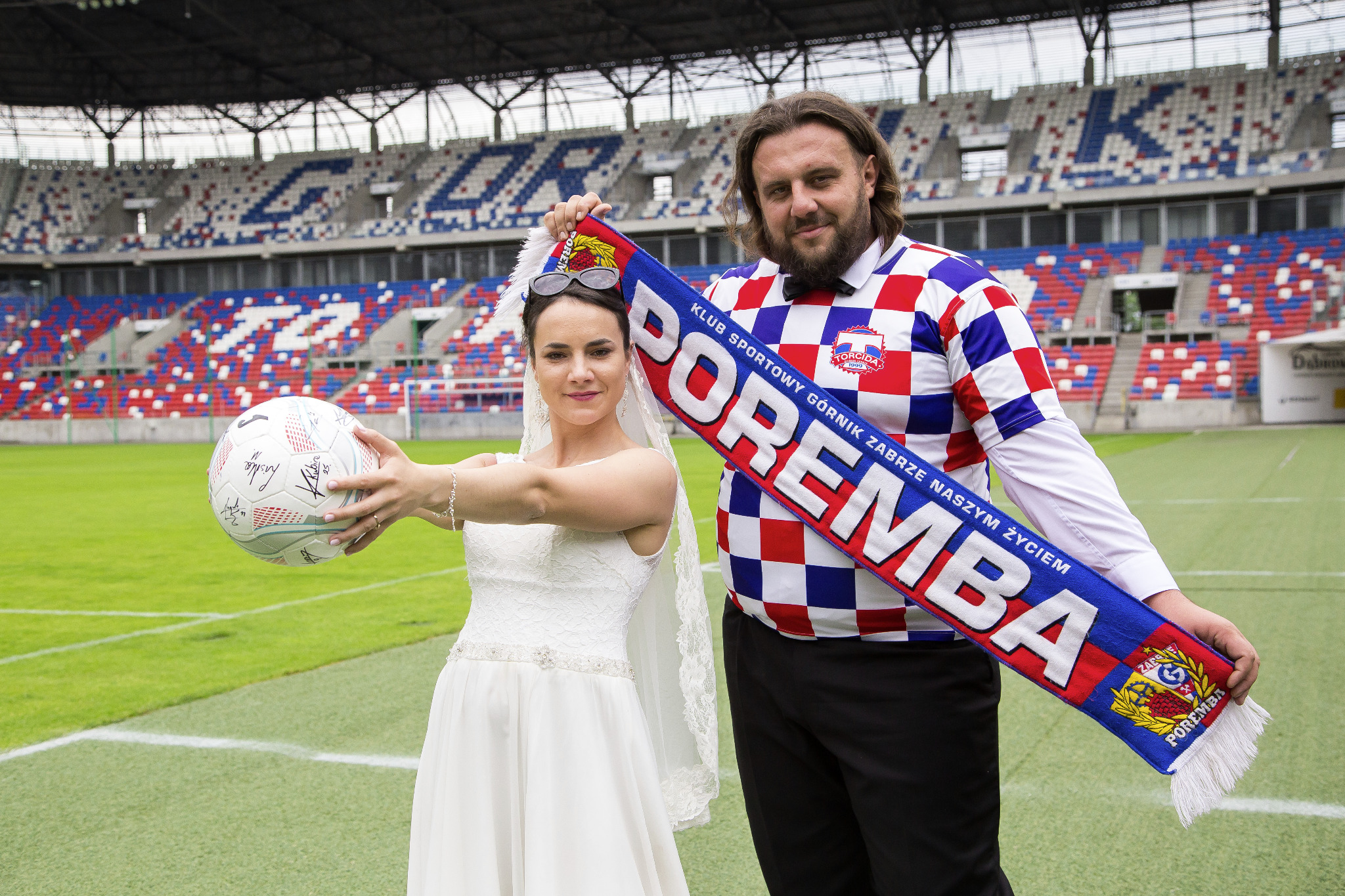 Para młoda na stadionie w Zabrzu: panna młoda w białej sukni trzyma piłkę z autografami, pan młody w koszuli w szachownicę i szaliku Górnika Zabrze.