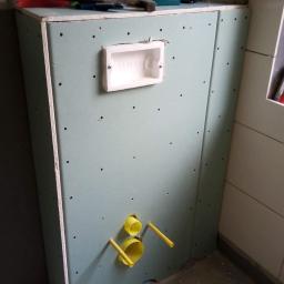 WZ. Wykończenia wnętrZ - Stelaż podtynkowy z płyt kartonowo-gipsowych w trakcie montażu w łazience, widoczne żółte rury instalacyjne, puszka Geberit, wkrętarka i narzędzia na górze konstrukcji.