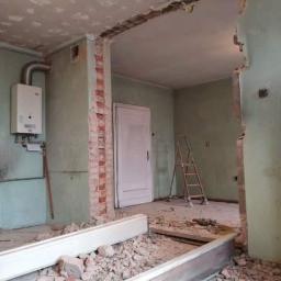 WZ. Wykończenia wnętrZ - Demontaż ściany działowej z odsłoniętą cegłą, widoczne gruz i elementy konstrukcyjne, w tle drabina oparta o ścianę i bojler gazowy na ścianie
