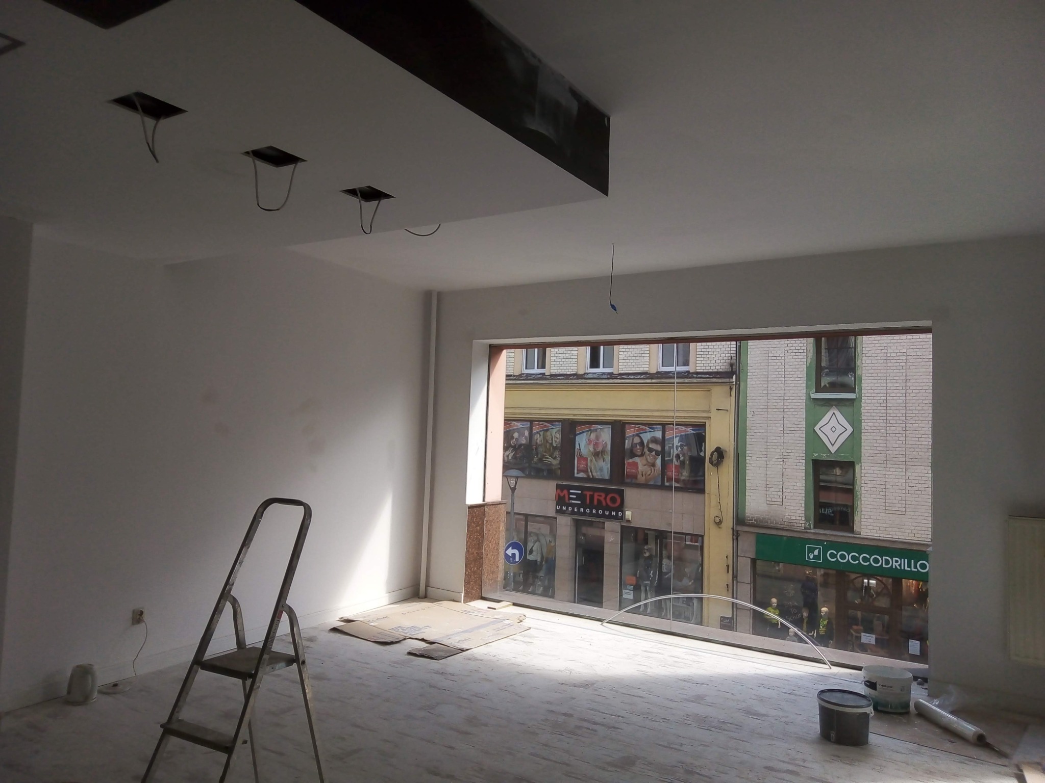 Wnętrze w trakcie malowania na biało, widoczna drabina, puszki z farbą i okno z widokiem na ulicę z sklepami Metro i Coccodrillo.