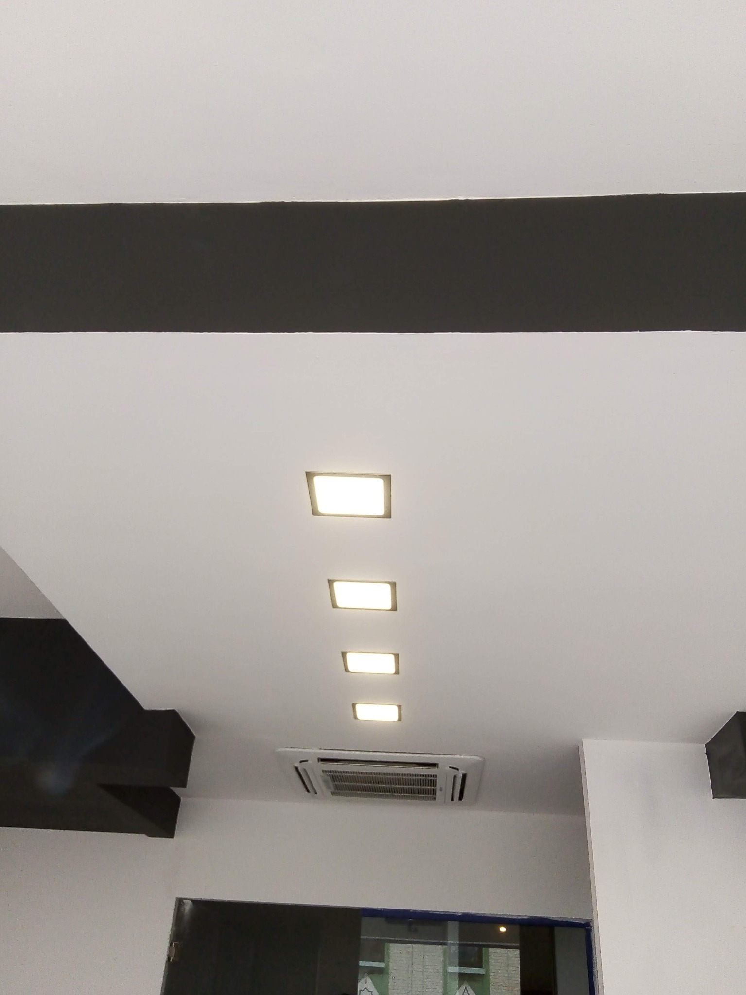 Biały sufit z rzędem wpuszczonych, kwadratowych lamp LED i klimatyzatorem, obramowany czarnymi elementami dekoracyjnymi.