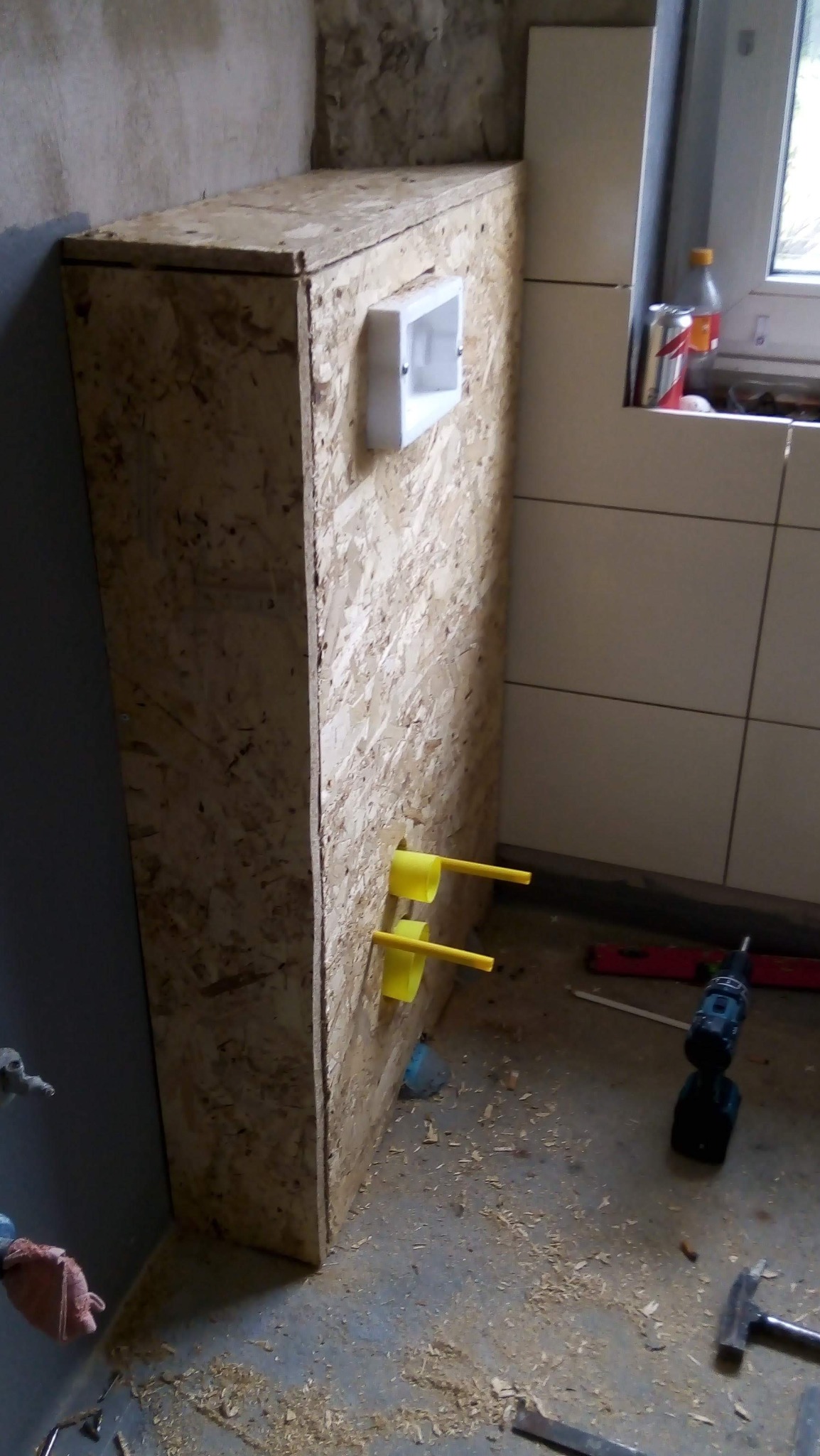 Konstrukcja z płyty OSB, prawdopodobnie obudowa stelaża WC, z widocznymi żółtymi rurami doprowadzającymi wodę i puszką elektryczną, w trakcie prac wykończeniowych łazienki.