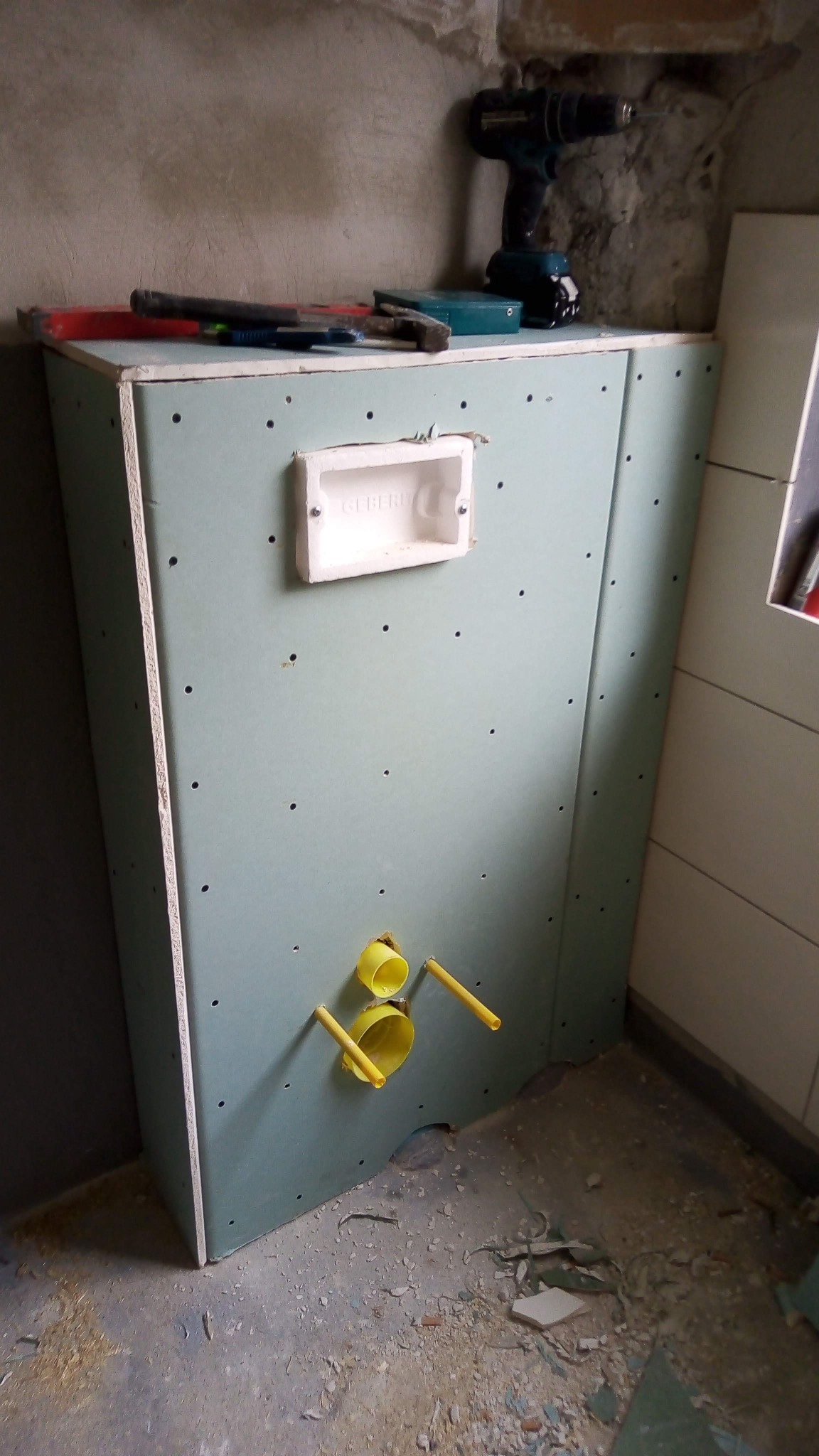 Stelaż podtynkowy z płyt kartonowo-gipsowych w trakcie montażu w łazience, widoczne żółte rury instalacyjne, puszka Geberit, wkrętarka i narzędzia na górze konstrukcji.