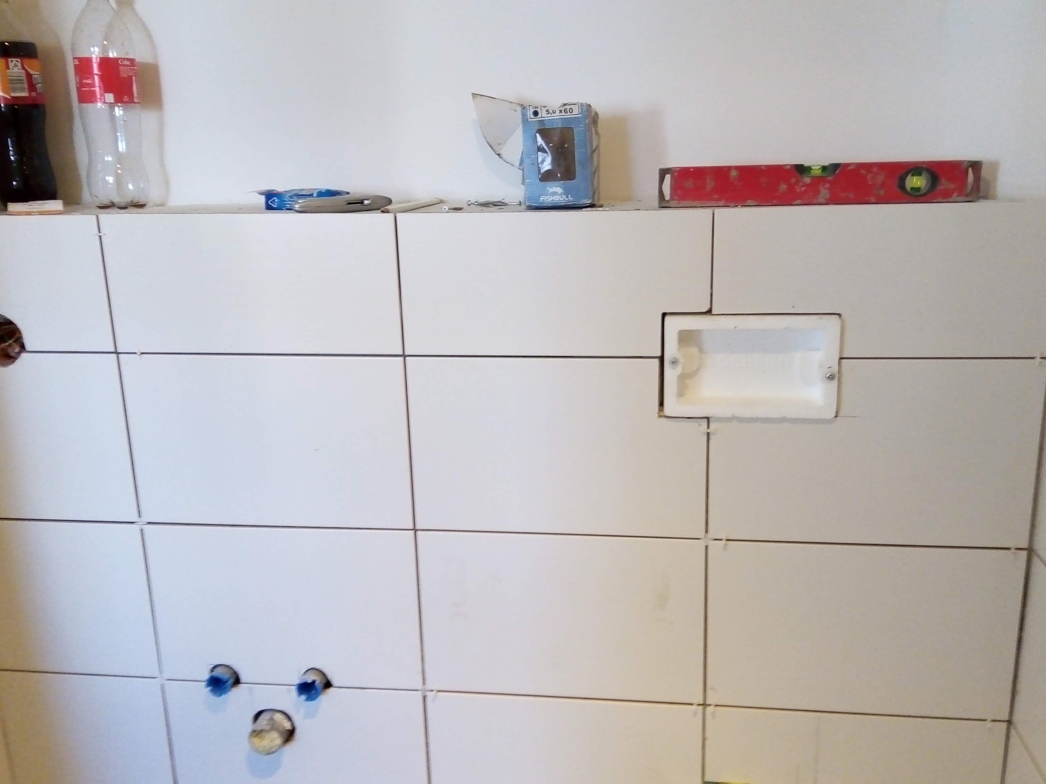 Ściana wyłożona białymi płytkami ceramicznymi, z widocznymi otworami na rury i puszką elektryczną, na górze poziomica, opakowanie po kołkach i butelki.