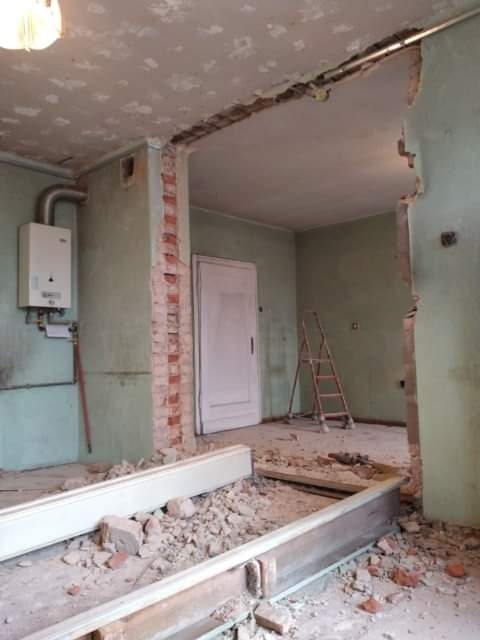 Demontaż ściany działowej z odsłoniętą cegłą, widoczne gruz i elementy konstrukcyjne, w tle drabina oparta o ścianę i bojler gazowy na ścianie