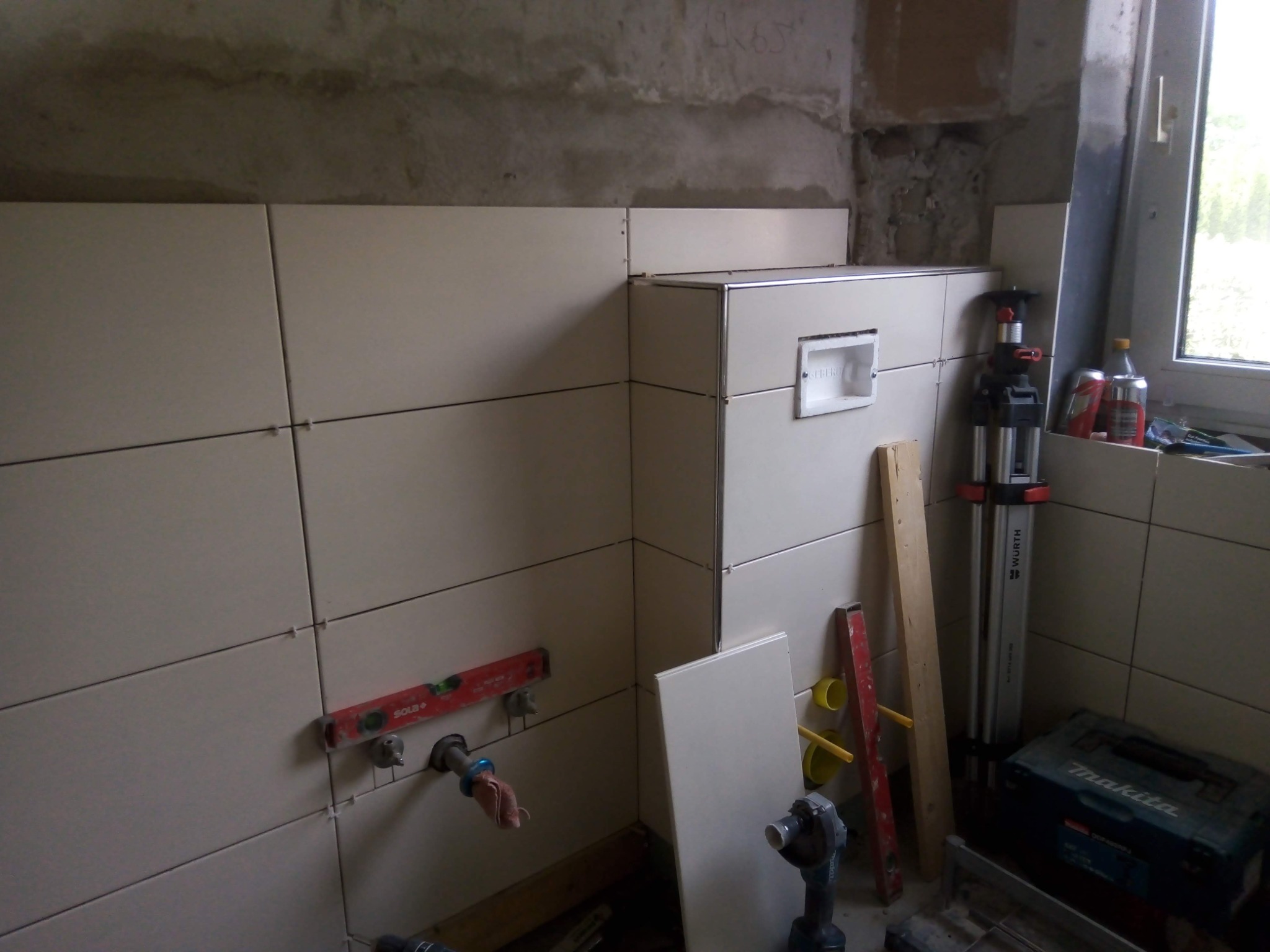 Montaż beżowych płytek ceramicznych w łazience, widoczne poziomice, laser krzyżowy Wurth, fragment instalacji wodnej i niezakończone fugowanie.