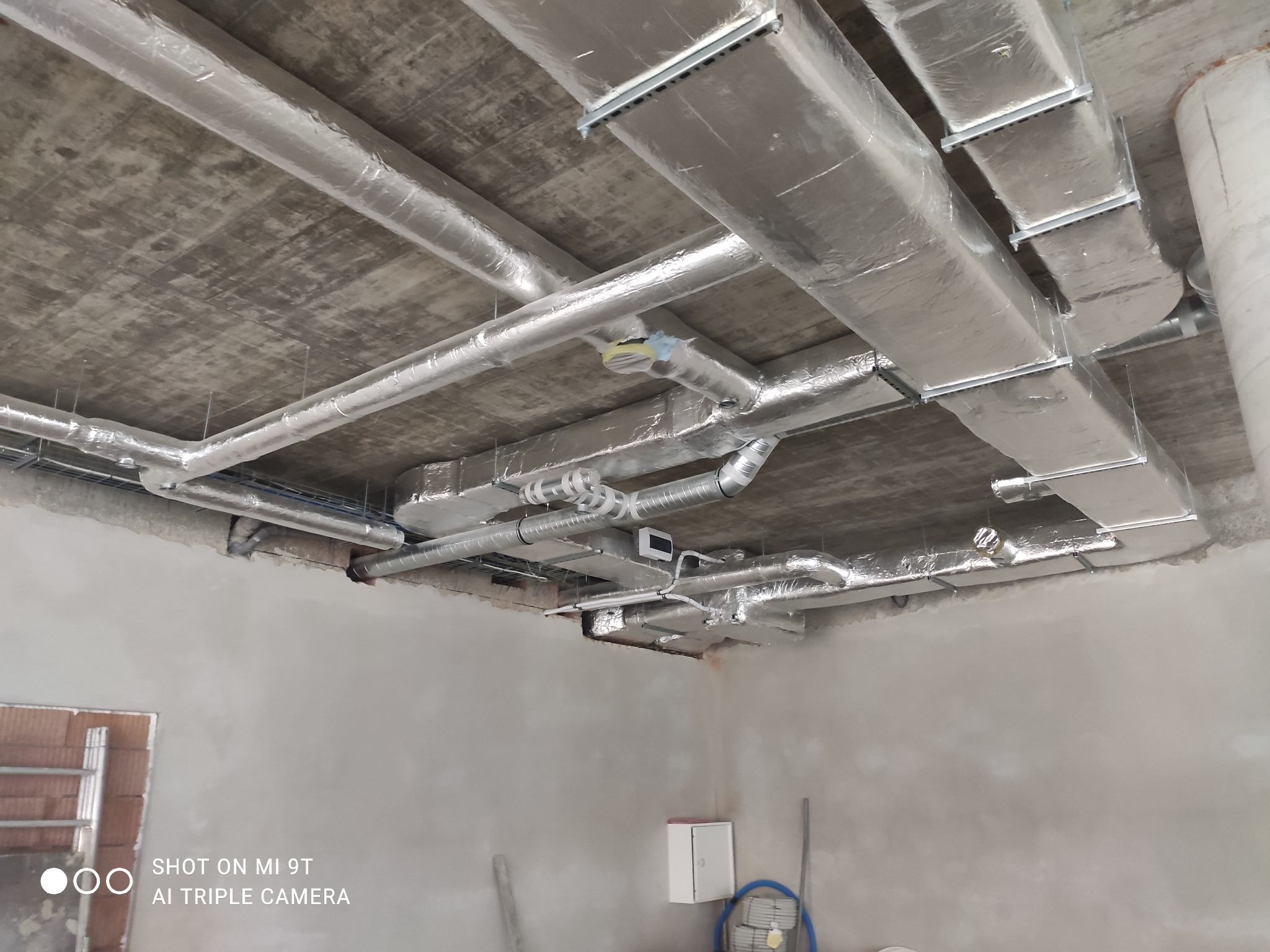 Widok pod sufitem w surowym stanie przedstawiający zamontowane, izolowane kanały wentylacyjne o przekroju prostokątnym i okrągłym, połączone kształtkami i zawieszone na metalowych profilach.