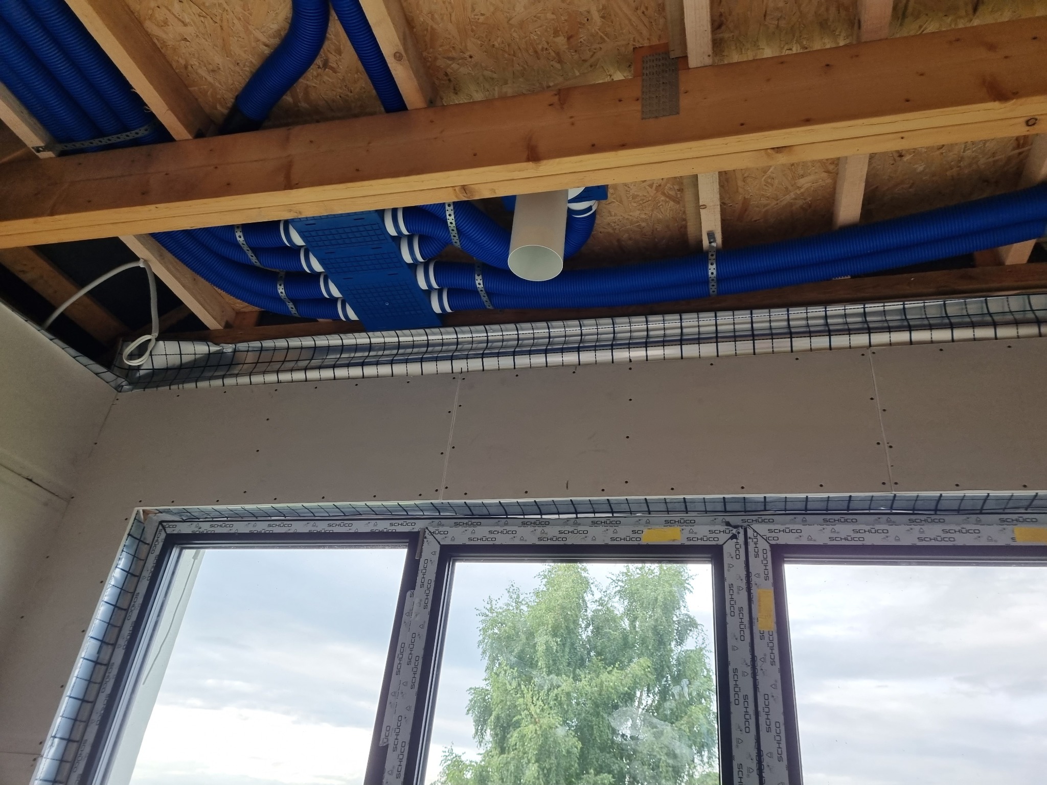 Montaż niebieskich rur wentylacyjnych pod stropem z desek, widoczne mocowania i elementy systemu wentylacji, okno z widokiem na drzewo w tle.