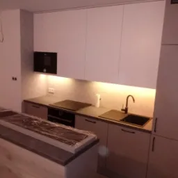 Nowoczesna kuchnia z białymi szafkami, czarną armaturą i blatem wyspowym zabezpieczonym folią ochronną, oświetlenie LED pod szafkami górnymi.