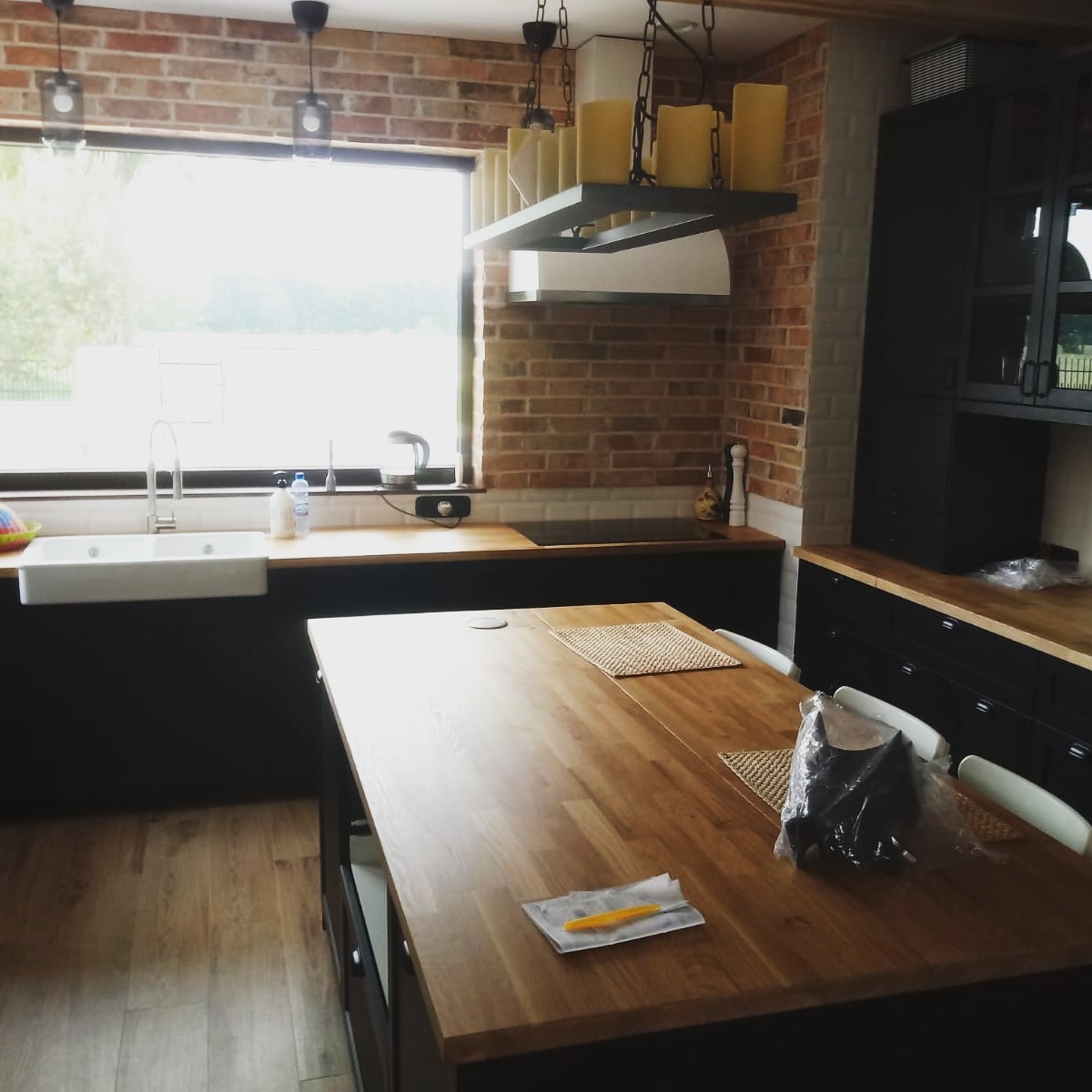 Kuchnia w stylu industrialnym z ceglaną ścianą, drewnianym blatem i wyspą kuchenną z widocznym oświetleniem wiszącym przypominającym świece.